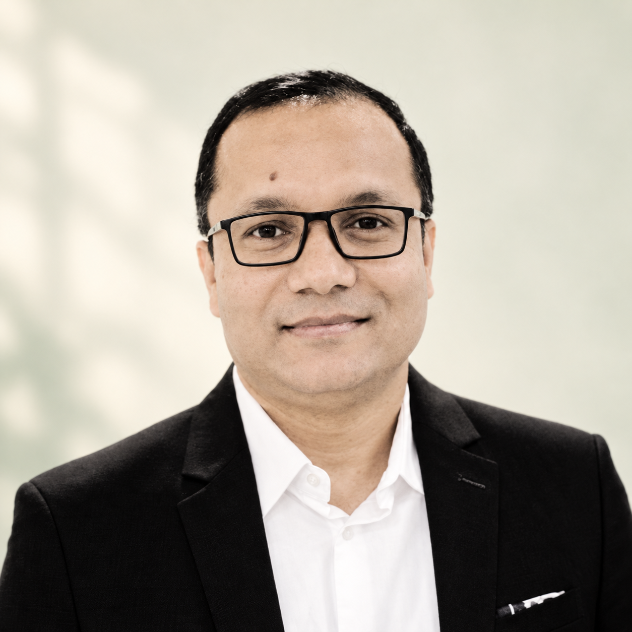 Portrait of Billal Hossain wearing glasses and a dark suit against a soft cream and sage background